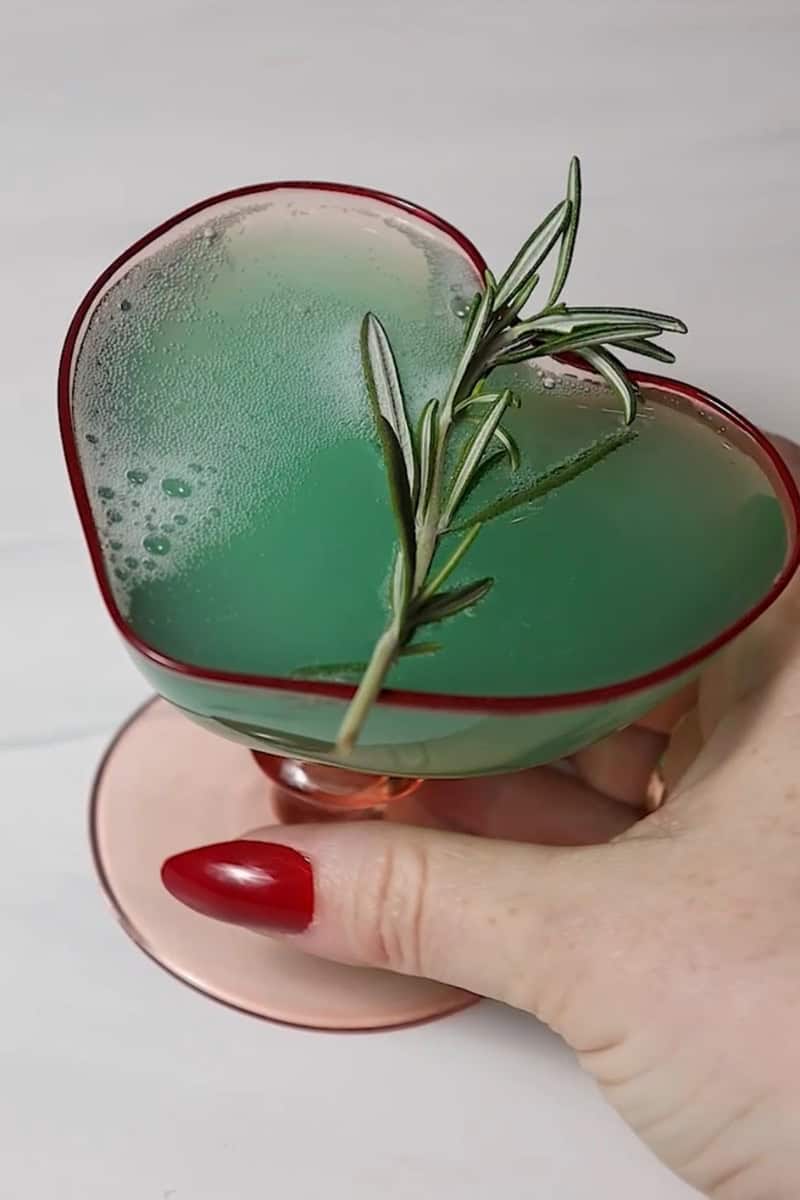 grinch drink in a heart shaped glass with a rosemary sprig