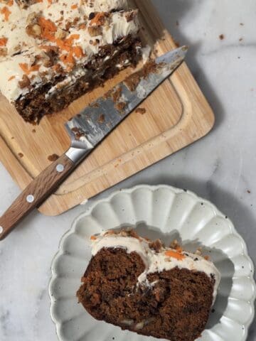close up of the baked and frosted protein carrot cake on a cutting board and a plate for serving.