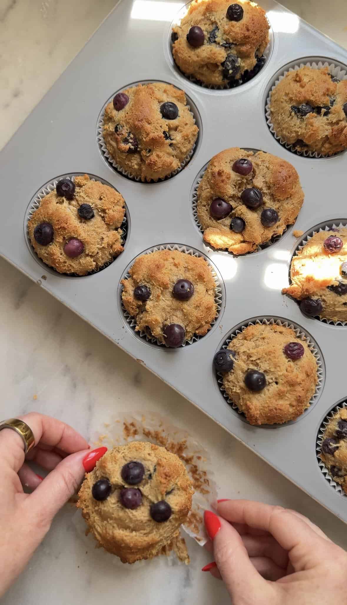 Finished Gluten Free Blueberry Cottage Cheese Protein Muffins in the tin, one being unwrapped from the parchment paper liner to show the texture of the muffin.
