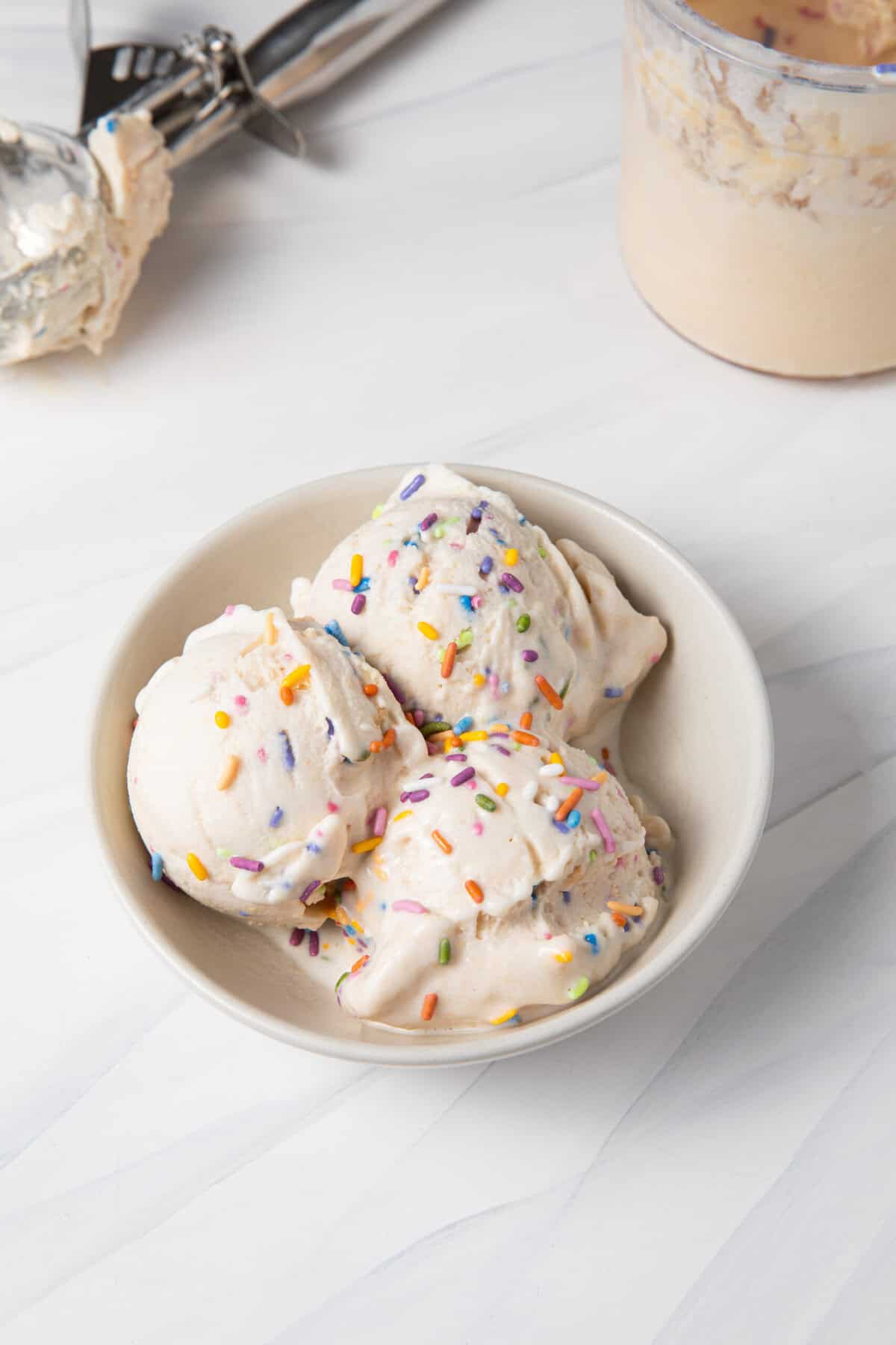 Birthday Cake Batter Protein Ninja Creami Ice Cream in a bowl, with the ice cream scoop and pint behind the bowl.