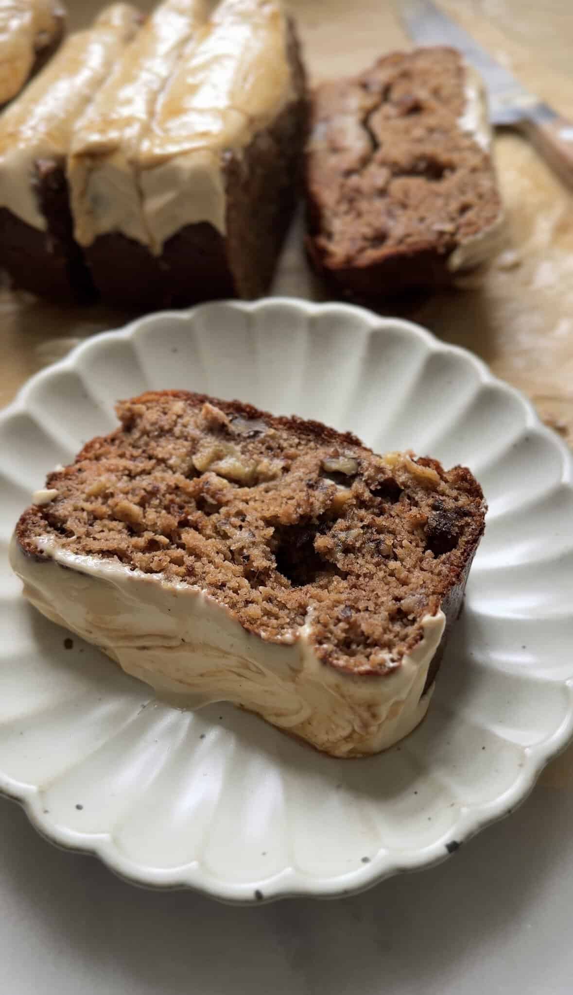 Slice of the Gluten Free Protein Banana Nut Bread on a plate with the loaf pictured behind it.