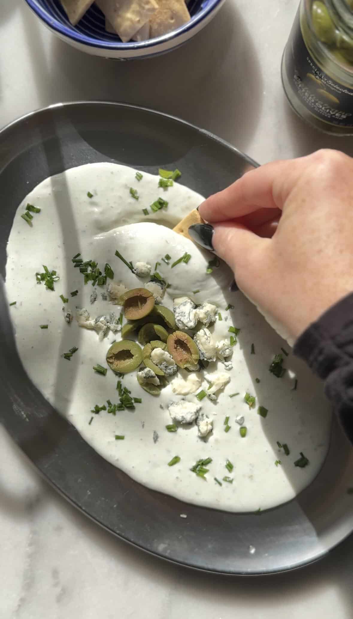 Dipping a gluten free cracker into the Protein Dirty Martini Dip topped with chives, olives, and blue cheese.